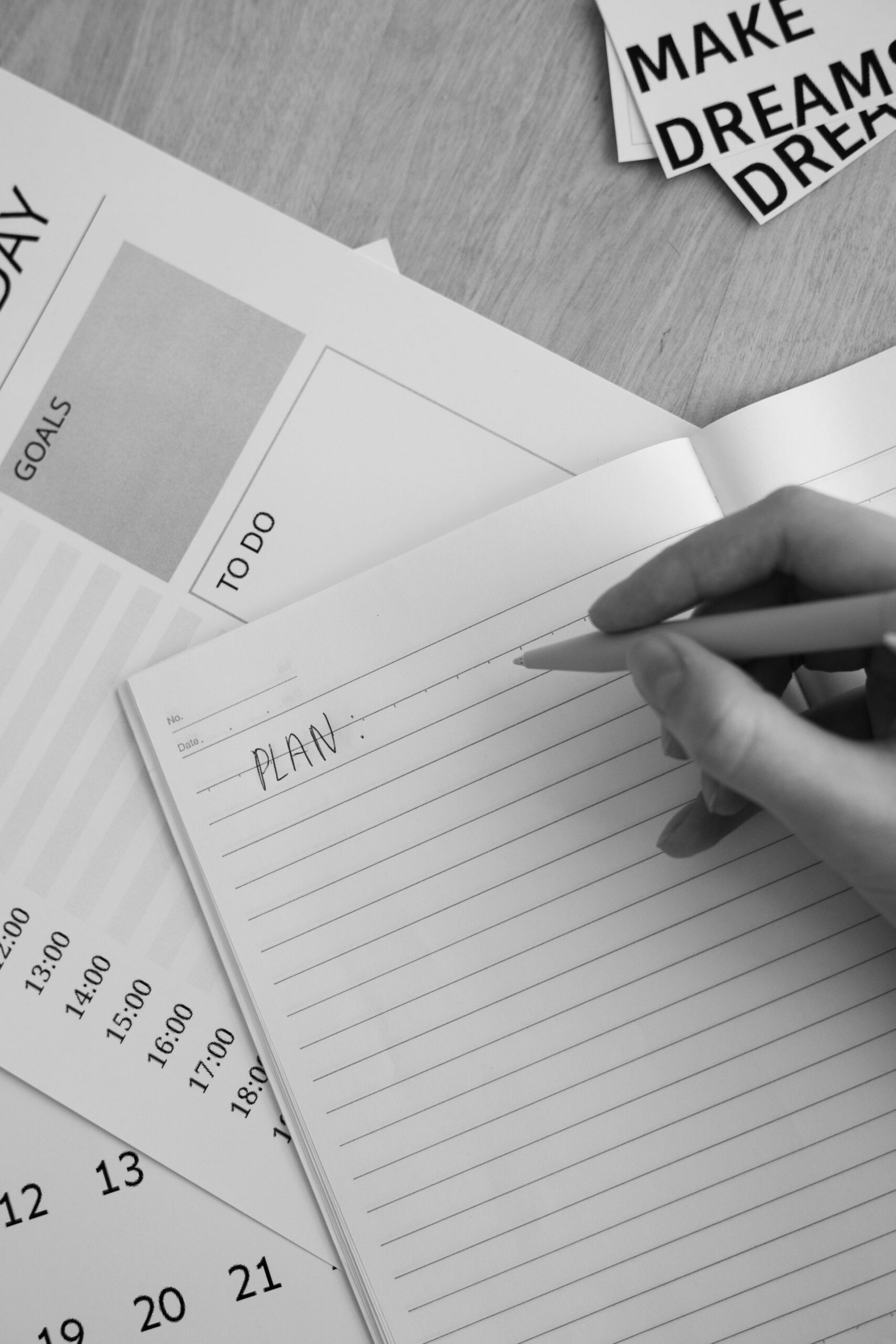 Black and white photo of hand writing 'plan' in notebook with planning materials around.
